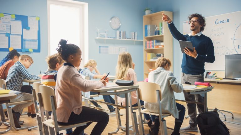 Enthusiastic Teacher Giving a Lesson in Elementary School to Class Full of of Diverse Children. Group of Smart Multiethnic Kids Learning New Stuff